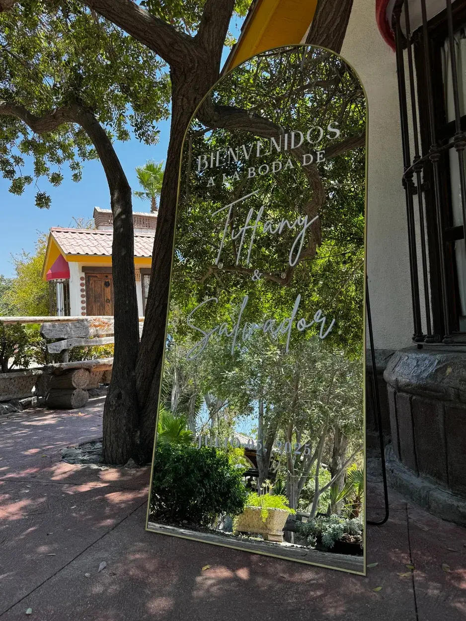 Espejo de bienvenida con texto blanco y parte superior curva, junto a un árbol en una boda al aire libre